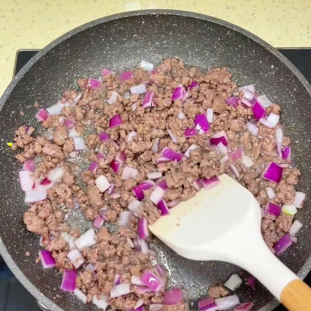 フライパンで炒められた挽肉と刻んだ赤玉ねぎにソースが加えられている様子。