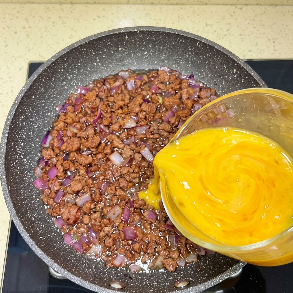 計量カップから、調理中の牛肉と玉ねぎが入ったフライパンに溶き卵が流し込まれている様子。