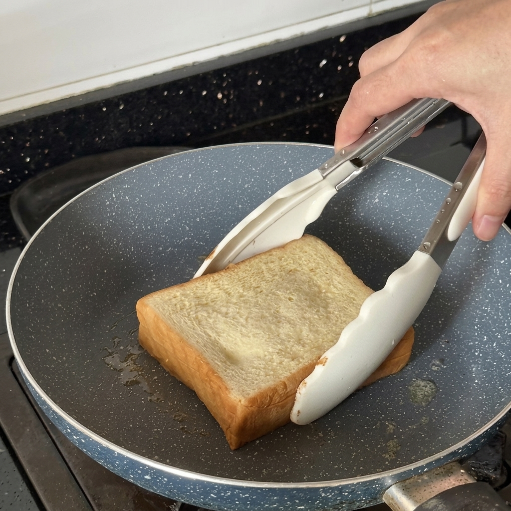 白いトングを使って、フライパンで厚切り食パンを焼いている様子。