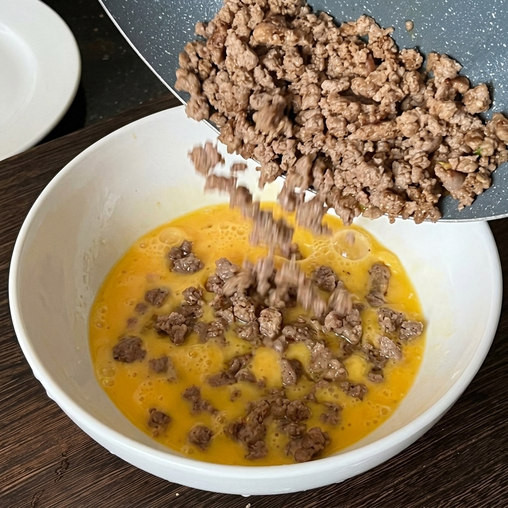 フライパンで炒めた牛ひき肉を、溶き卵の入ったボウルに注ぎ入れている様子。