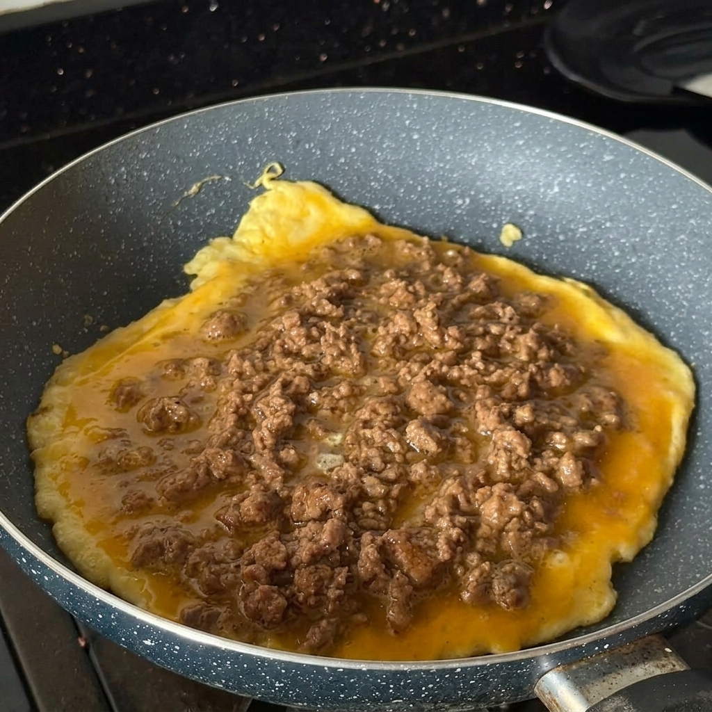 斑点模様のフライパンで丸いオムレツを焼き、その上に味付けした牛ひき肉をたっぷりと乗せている様子。