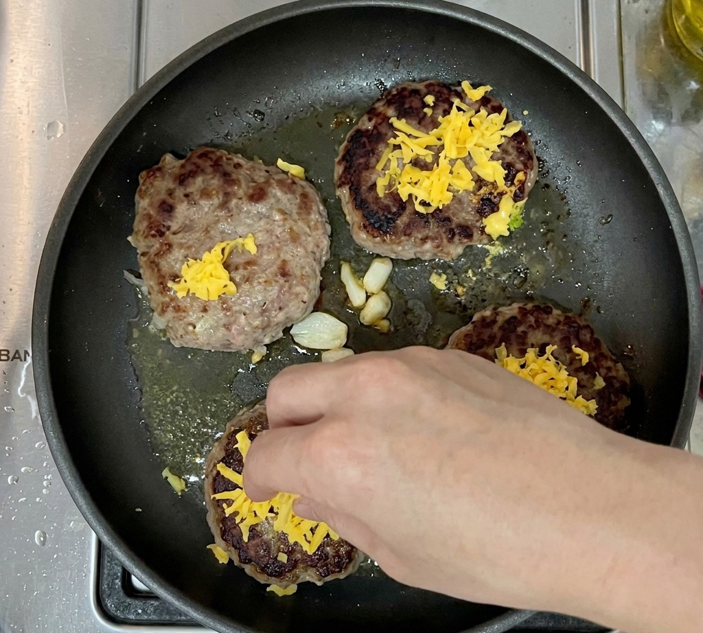 フライパンの中で、チェダーチーズの山がトッピングされた4つの焼きあがったパティ。
