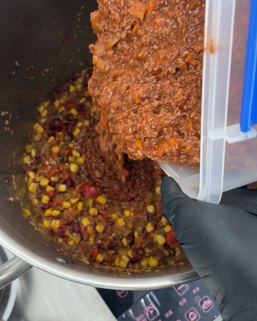スパイスと野菜が入った大きめの鍋に、濃厚なミートソースを注ぎ入れている様子。