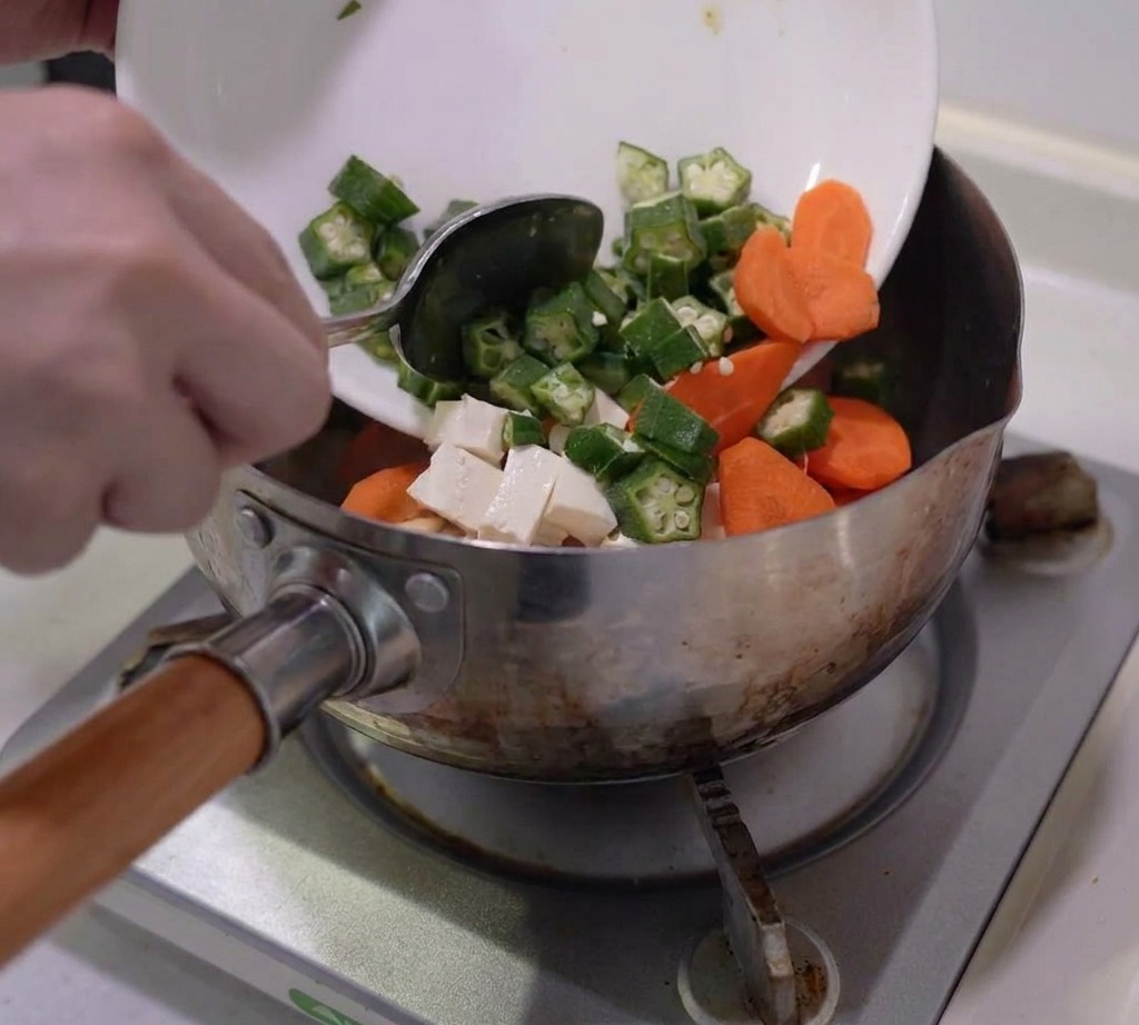 下準備したオクラ、にんじん、しめじ、豆腐をステンレスの鍋に入れる様子。