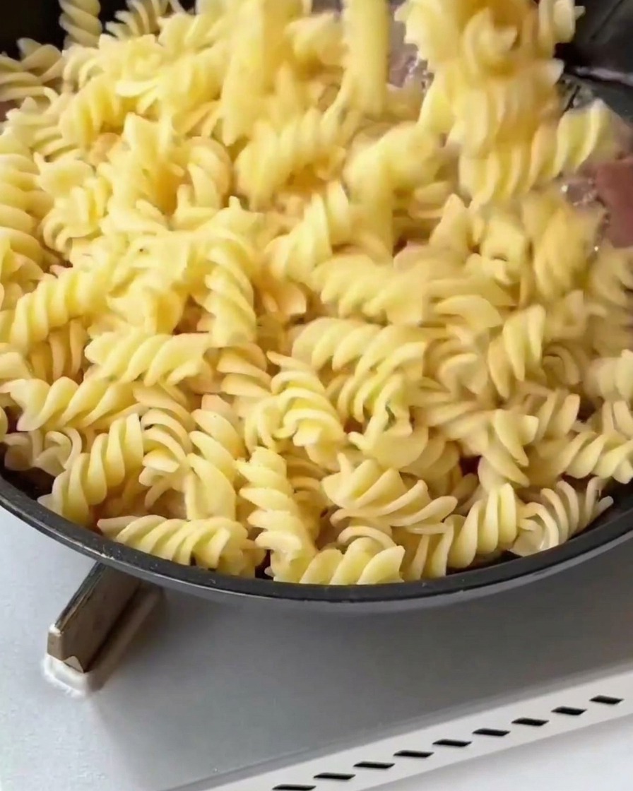 茹でたフジッリとアスパラガスを、焼いたベーコンと玉ねぎが入ったフライパンに加えている様子。