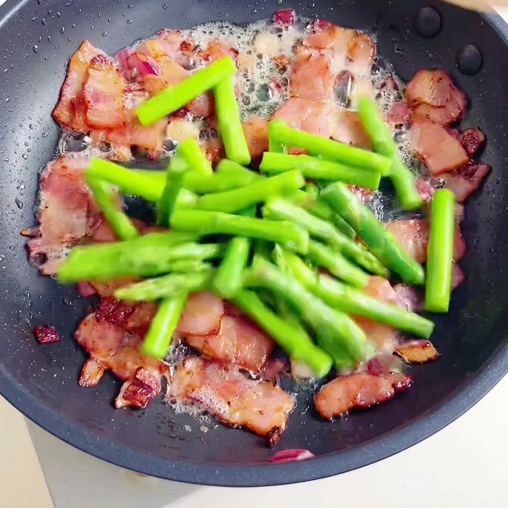 カリカリのベーコンにパスタとアスパラを加えて合わせる様子。