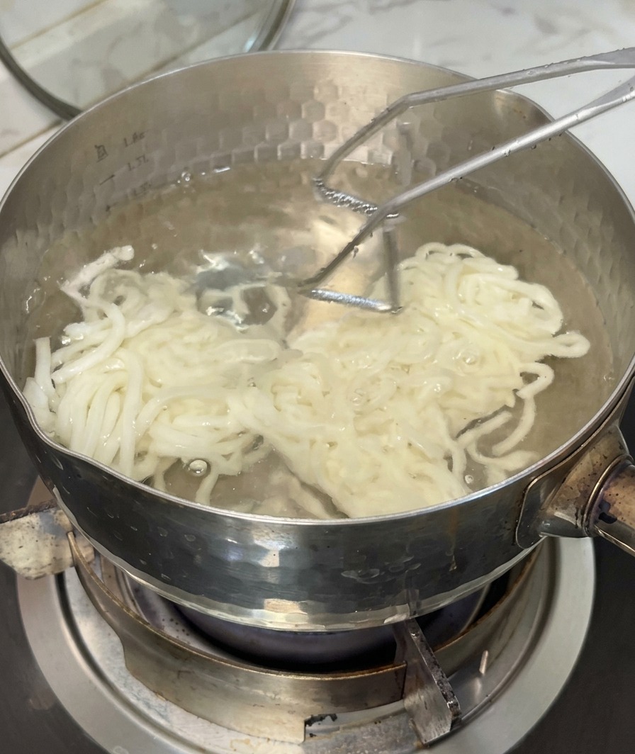 沸騰した鍋から、金属製のザルでモチモチのうどんをすくい上げている様子。