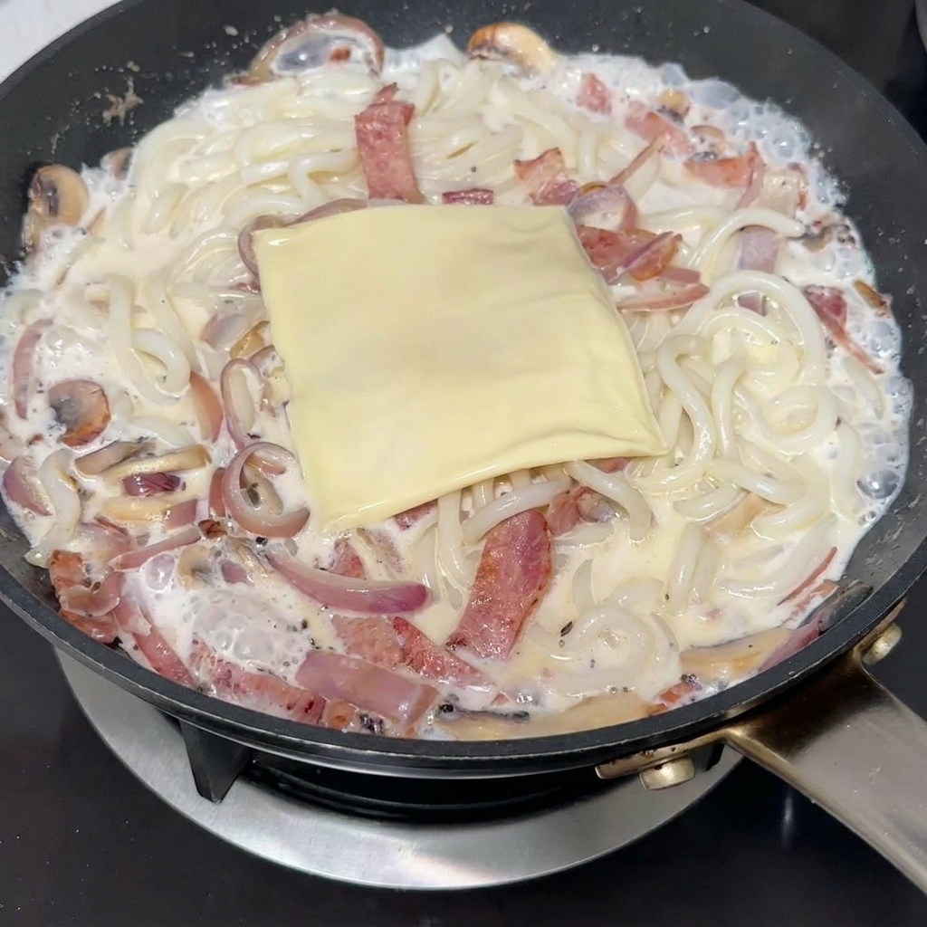 フライパンの中のクリーミーなうどんと具材の上に、四角いスライスチーズが乗っている様子。