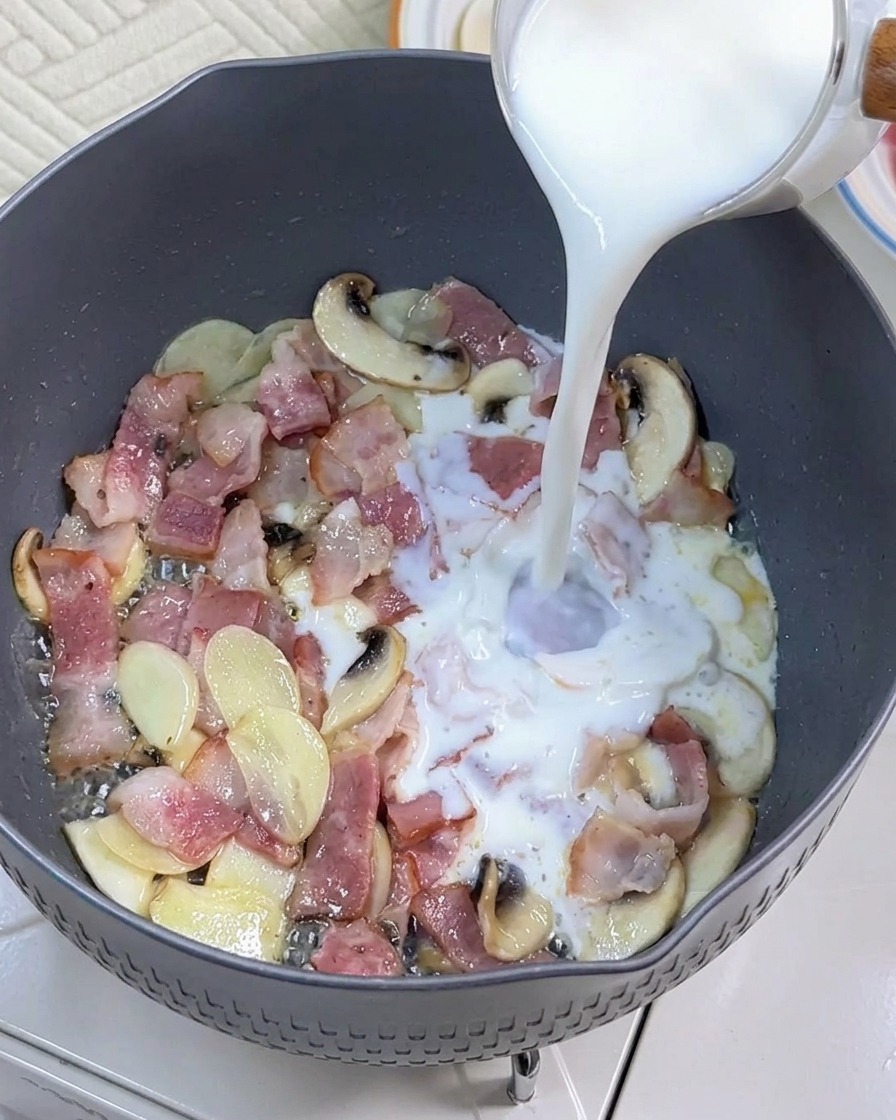 炒めたベーコンときのこのフライパンに牛乳を注いでいる様子。