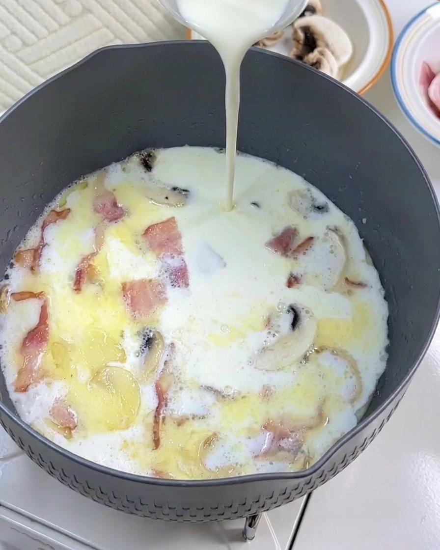 牛乳、ベーコン、きのこが入ったフライパンに生クリームを加えている様子。