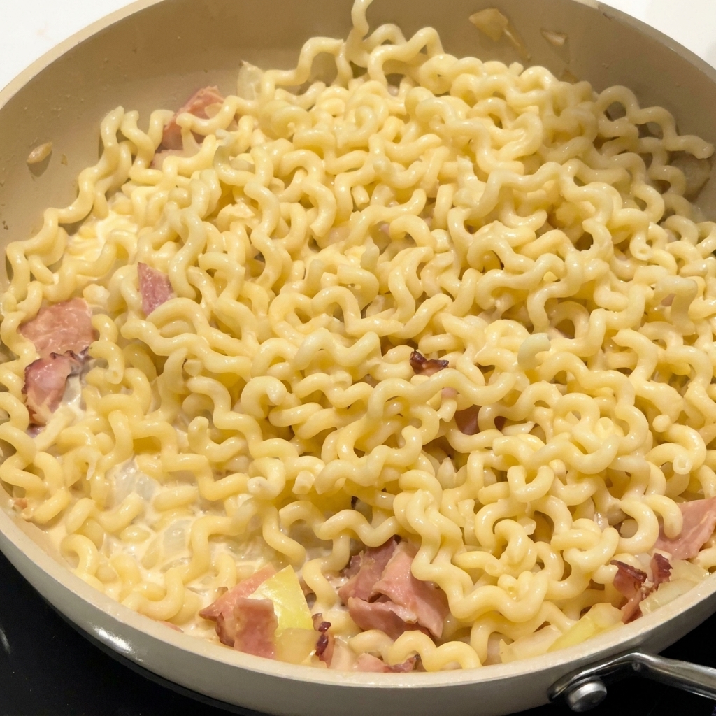 フライパンの中でクリーミーなソースに絡んだカールパスタとベーコンに調味料を振りかけている様子。