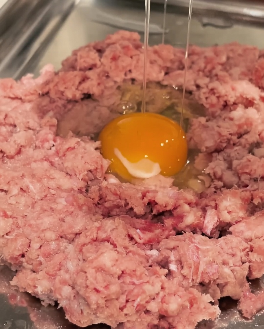 ボウルに入った牛ひき肉に生卵を割り入れる様子。