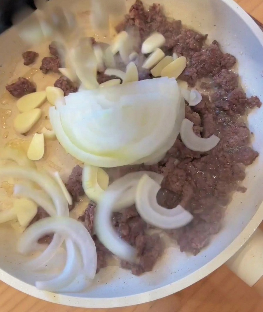 炒めたひき肉のフライパンに、スライスした玉ねぎとにんにくを加えている様子。