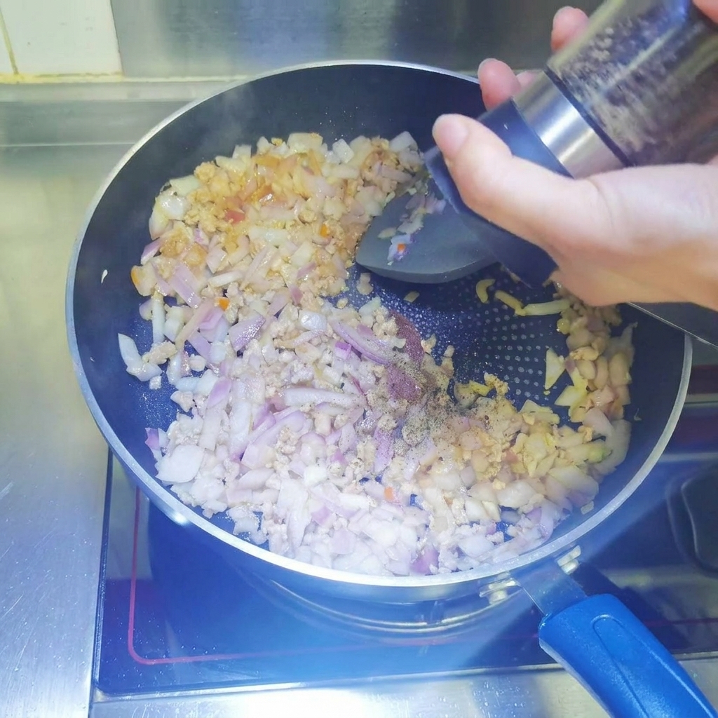 ひき肉と角切り玉ねぎが入ったフライパンに、ペッパーミルで黒こしょうを挽き入れている様子。