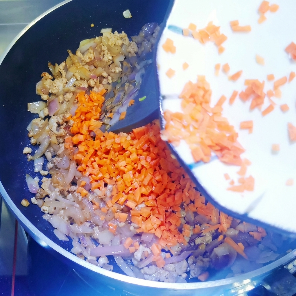 炒めた肉と玉ねぎの入ったフライパンに角切りのにんじんを加え、シリコンスパチュラで混ぜ合わせている様子。