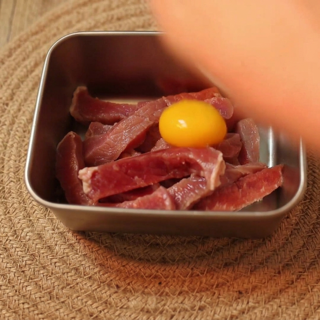 金属製のボウルに入ったスライス牛肉の上に、鮮やかな黄色の卵黄がのっている様子。