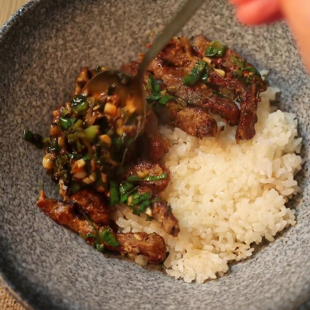 スプーンで熱々の牛肉とねぎダレを、白いご飯の上にたっぷり盛り付けている様子。
