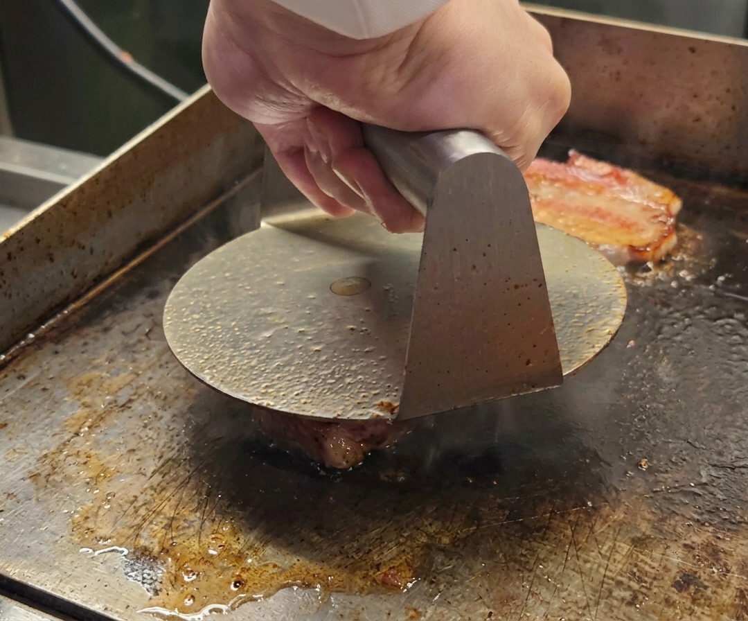 熱い鉄板の上で和牛ひき肉の塊を押し潰す金属製のバーガープレス。