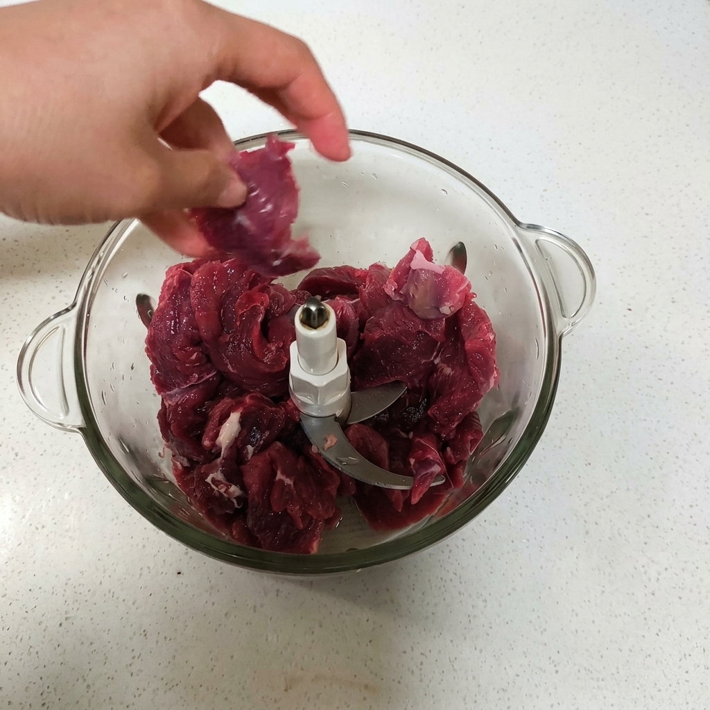 ガラス製のフードプロセッサーのボウルに、生の赤い牛肉の切り身を入れている手元の様子。