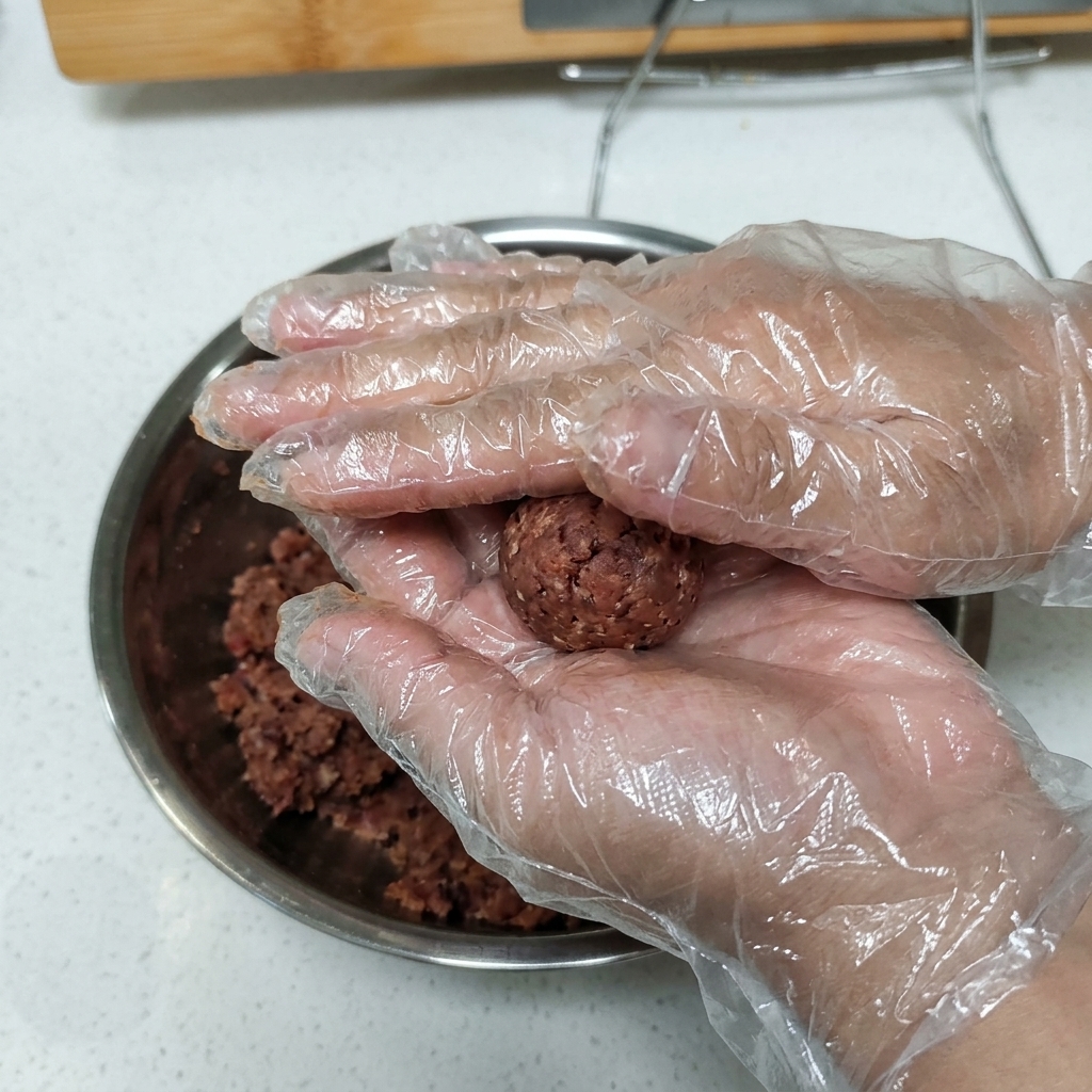 手袋をした手で、味付けしたひき肉のボールを持ち、パティの形に押し固めている様子。