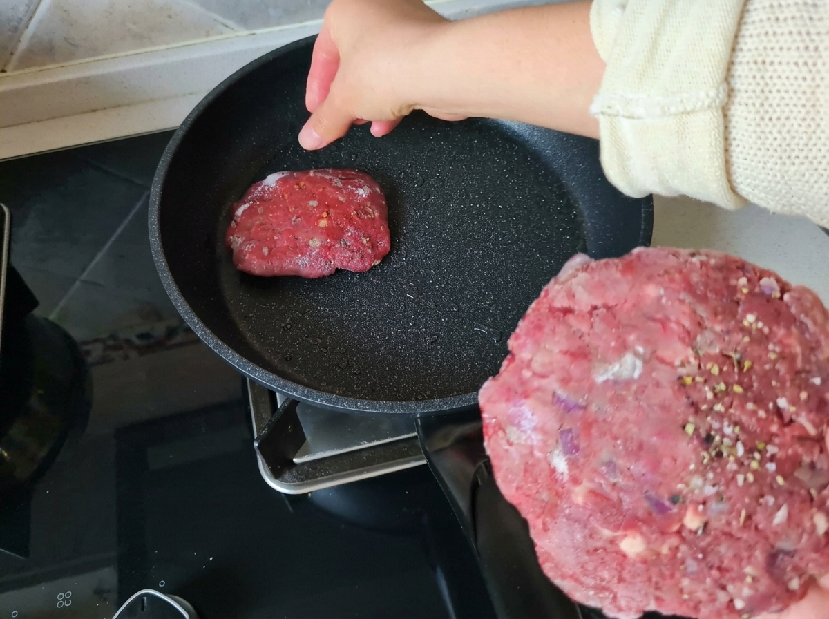 味付けされた生の丸いビーフパティを手に持ち、軽く油をひいた熱いフライパンに置いている様子。
