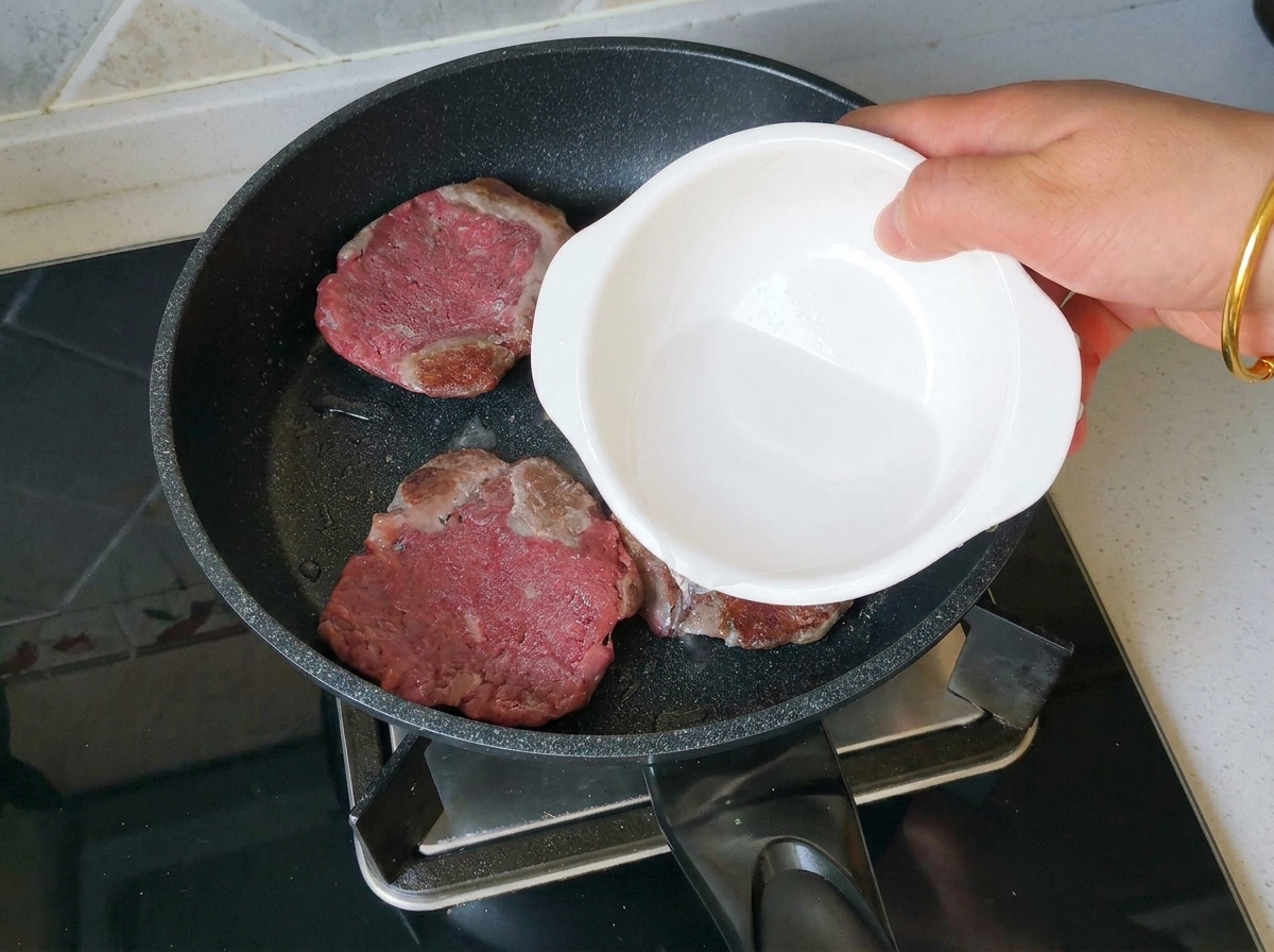 小さな白いボウルから、こんがりと焼けた3枚のビーフパティが入ったフライパンに少量の水を注いでいる手元の様子。