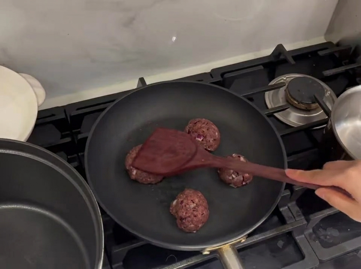 熱いフライパンの上で、金属のヘラを使って生の牛肉のボールを押しつぶして焼いている様子。