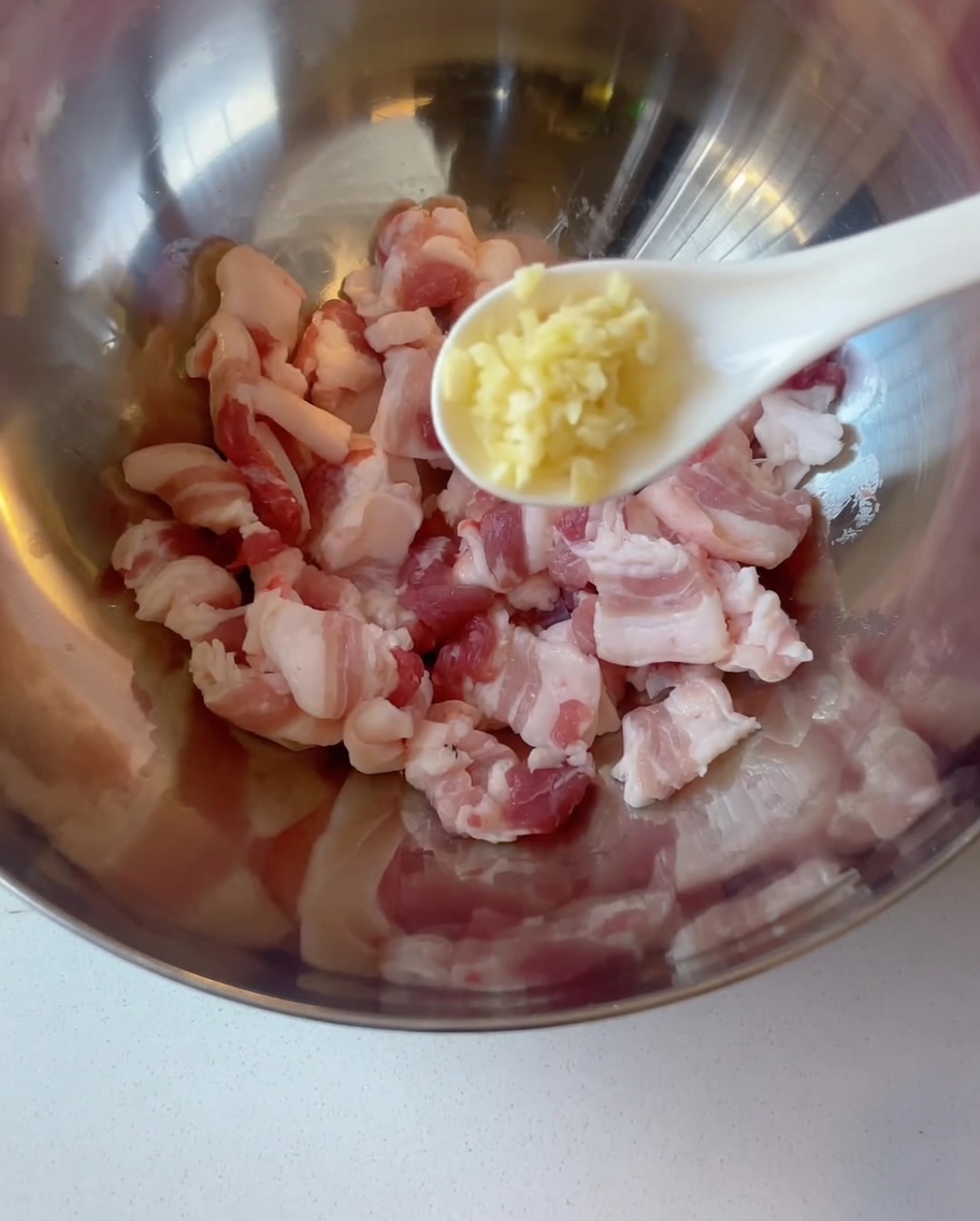 金属製のボウルに入った豚バラ肉に、スプーンでみじん切りのにんにくを加えている様子。