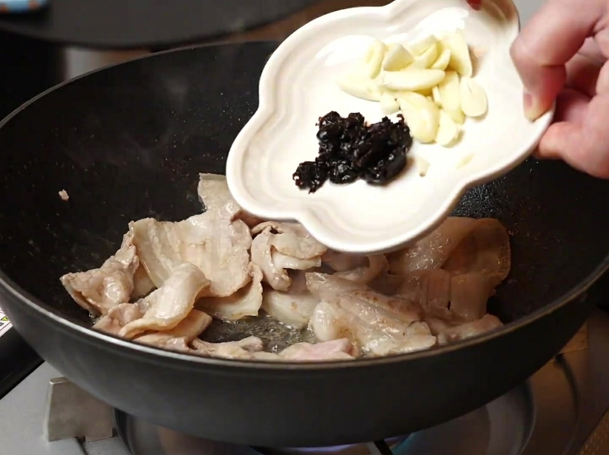 焼けた豚肉のフライパンににんにくとトウチを加える様子。