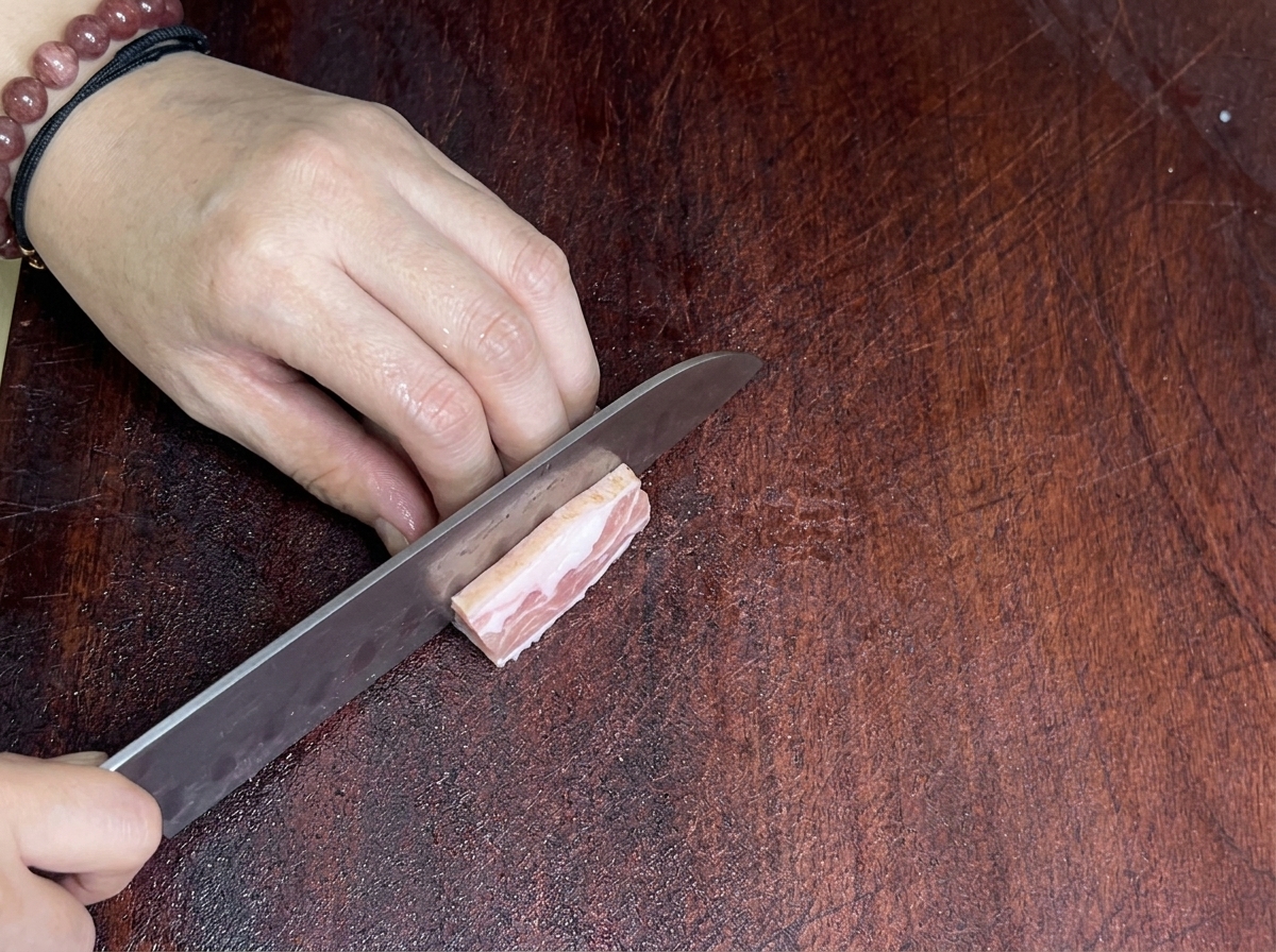 まな板の上で鋭利な包丁を使って豚バラ肉を薄切りにしている様子。