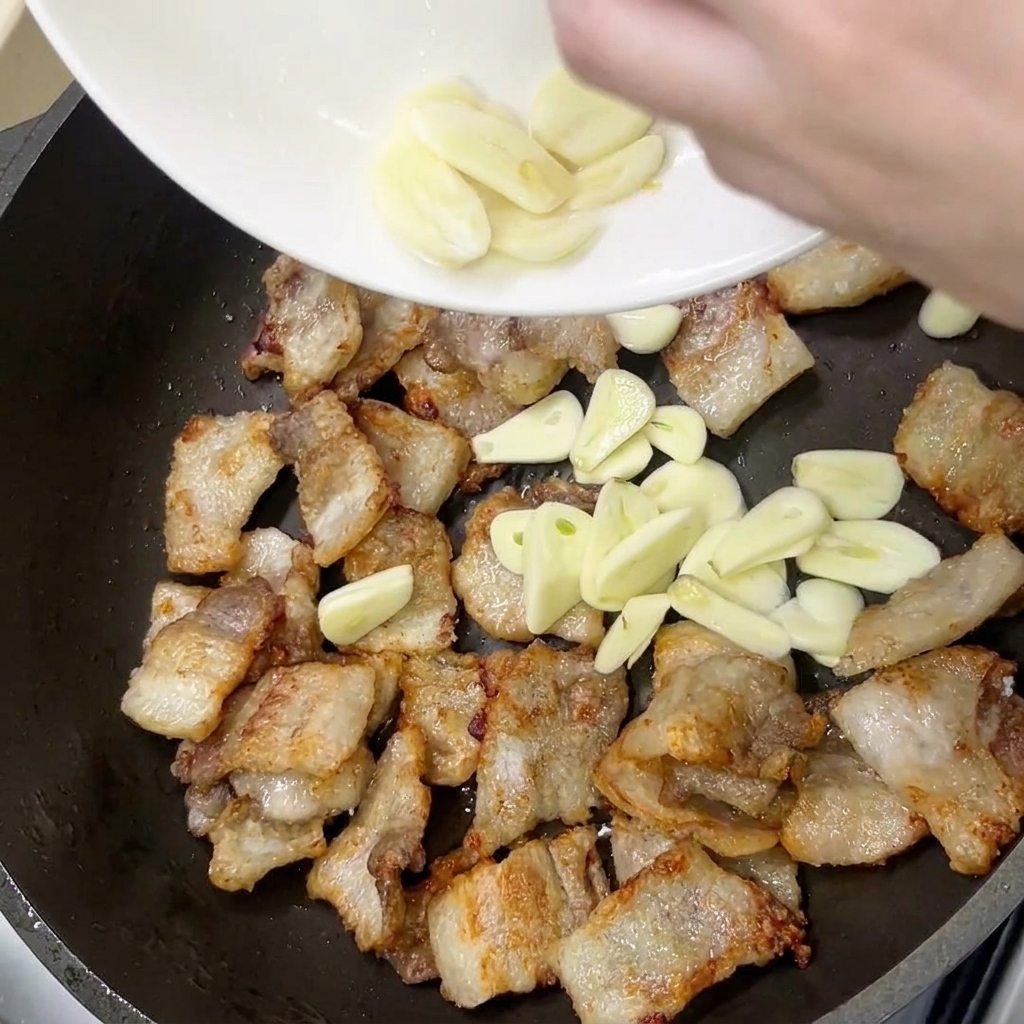 黄金色に焼けた豚バラ肉の隣で、スライスしたニンニクを炒めている様子。