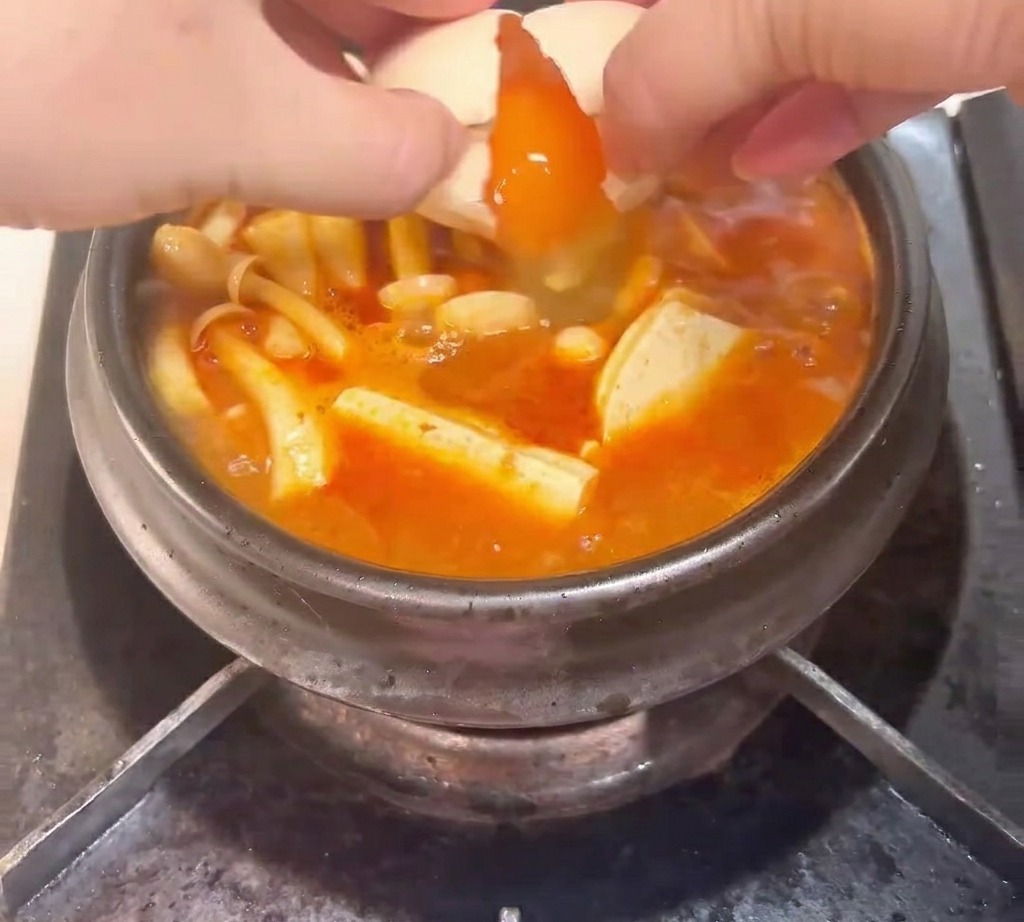 キムチ、ツナ、きのこ、豆腐が入ったぐつぐつ煮え立つ土鍋に、生卵を割り入れている様子。