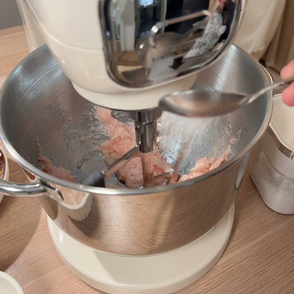 ミキサーボウルのひき肉にスプーンで調味料を加えている様子。周辺には他の材料が並んでいる。