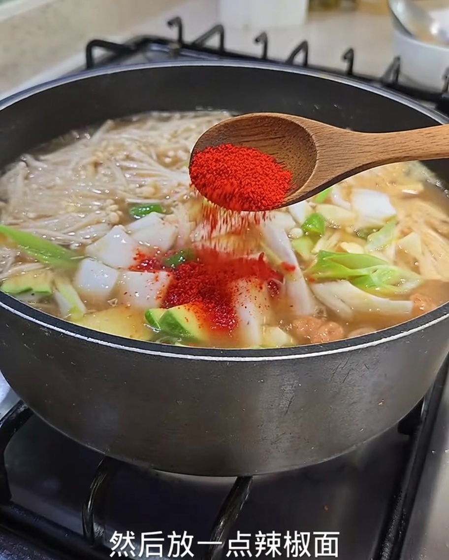ズッキーニ、玉ねぎ、えのき茸が煮えるチゲに、木べらから鮮やかな赤い粉唐辛子を振りかけているクローズアップ。