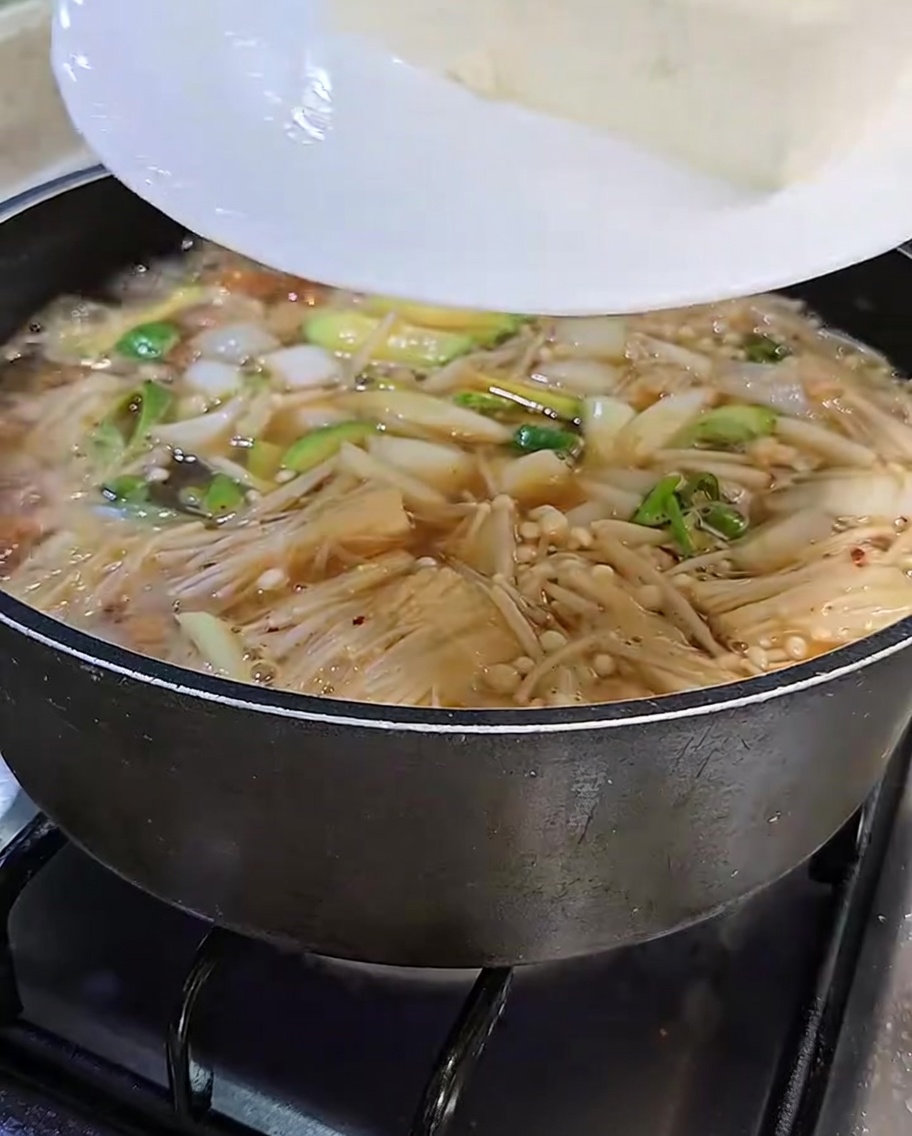 きれいにスライスされた白い豆腐を、風味豊かな味噌チゲの鍋に白いお皿からそっと移し入れている様子。