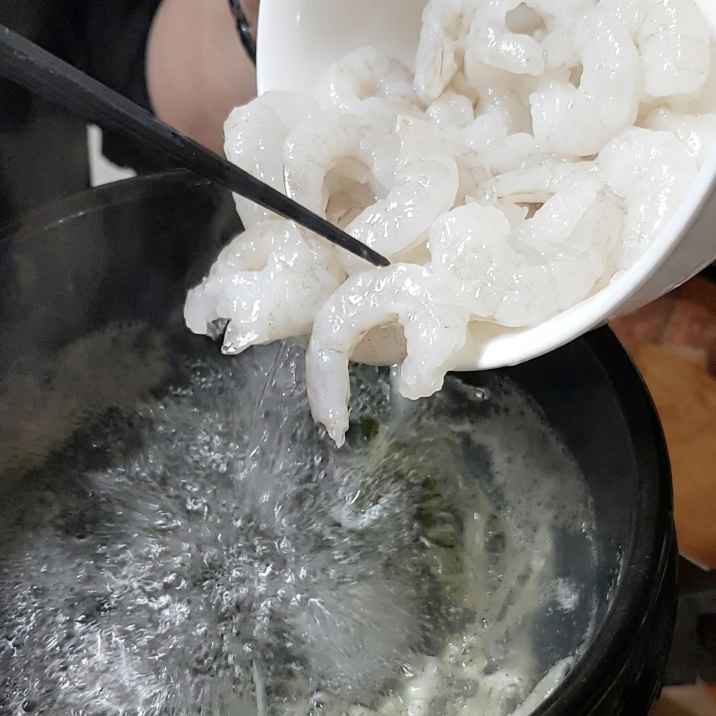 白いボウルから新鮮なエビをグツグツ煮立つ石鍋のスープに注ぎ入れている様子。