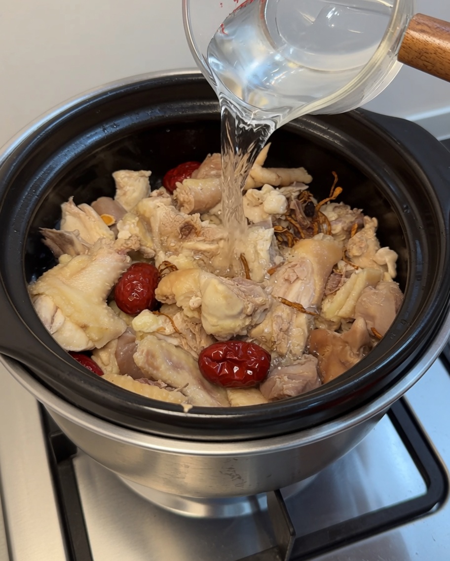鶏肉、なつめ、生薬が入った土鍋に、計量カップからお湯を注いでいる様子。