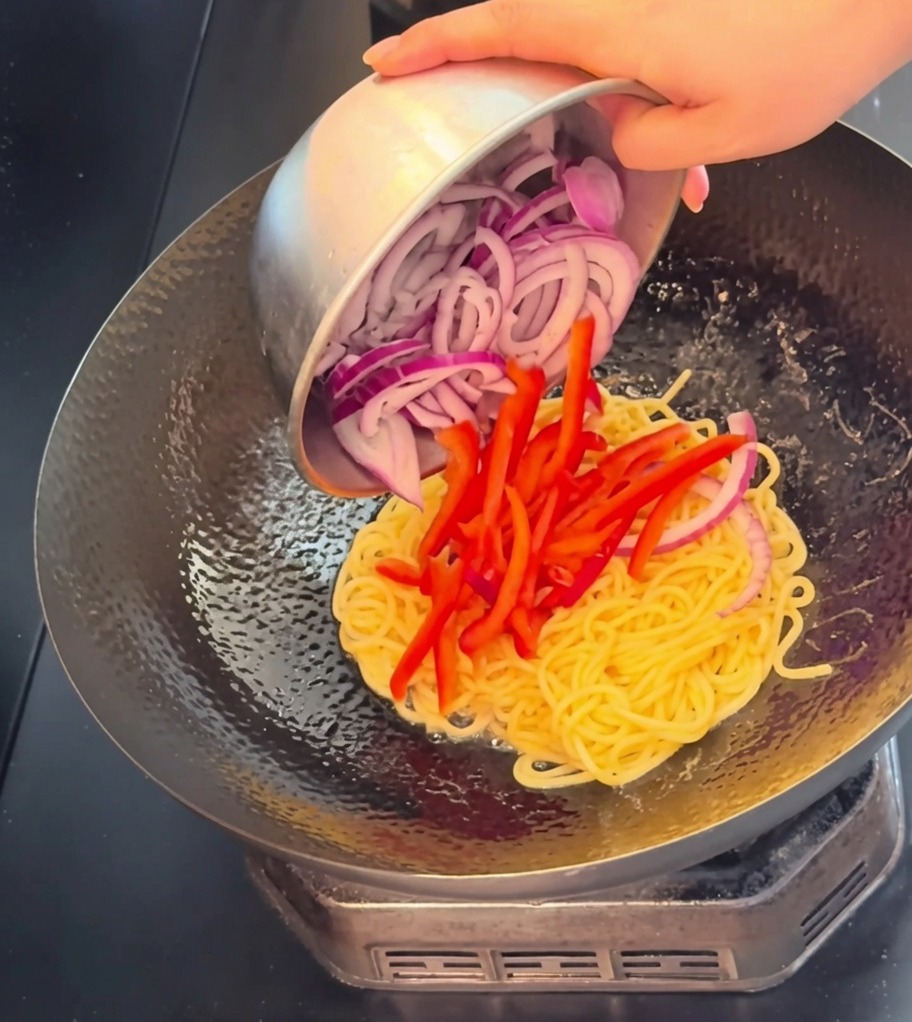 茹でたスパゲッティの入った中華鍋に、牛肉の薄切り、鮮やかな赤パプリカ、紫玉ねぎのスライスを加えているクローズアップ画像。