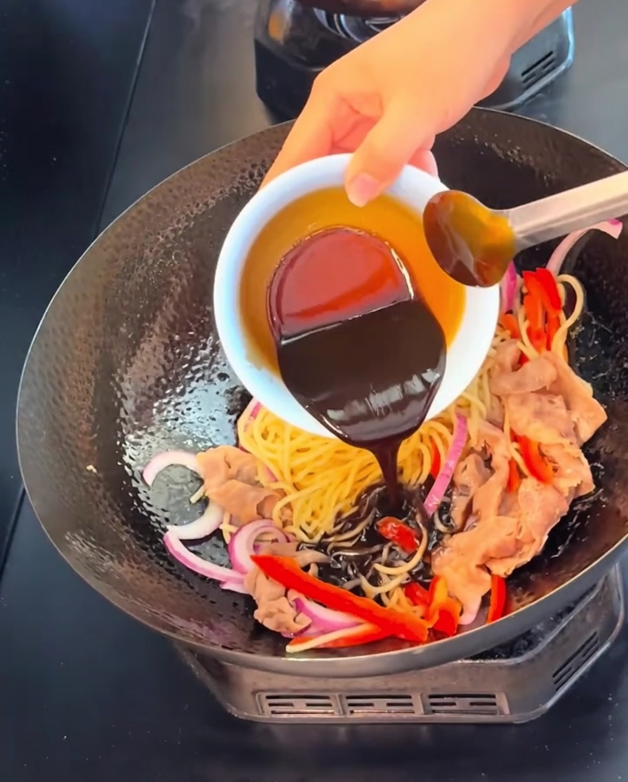 小さな白いボウルから、中華鍋の中の麺、牛肉、野菜の上に、濃厚で色濃い黒胡椒ソースをかけている様子。