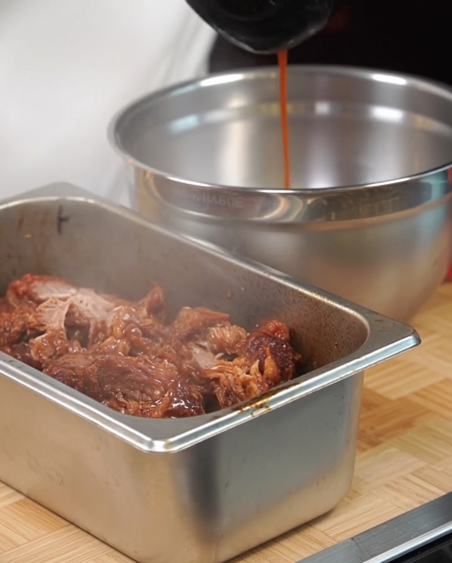 金属製のトレーに入ったほぐした豚肉に、煮汁をかけている様子。