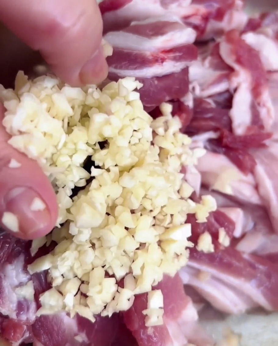 スライスした豚肉を入れた金属製のボウルに、みじん切りのニンニクを加えている様子。