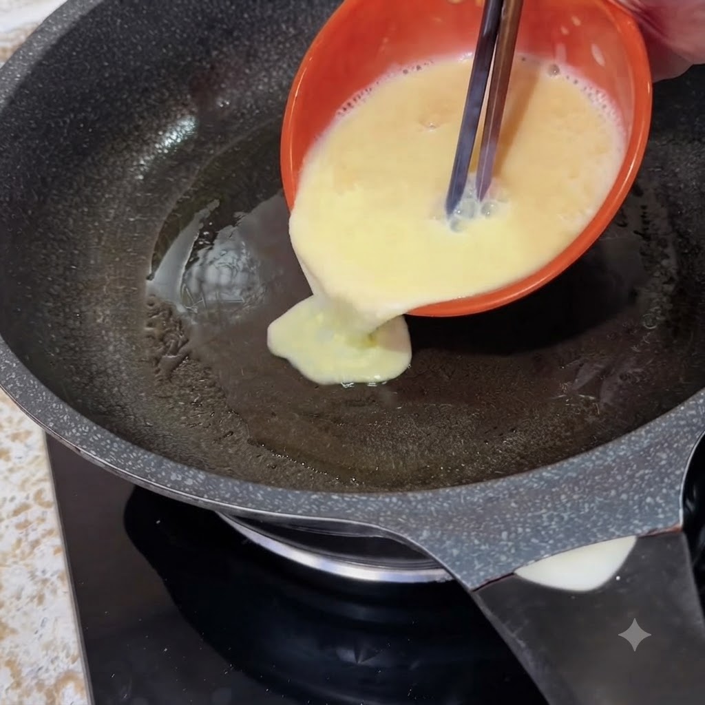 オレンジ色のボウルから、箸を使って溶き卵と牛乳の混合物を熱したフライパンに注いでいる様子。