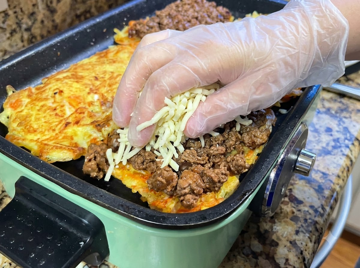 フライパンで焼いたポテトパンケーキにミートソースを広げ、その上からシュレッドモッツァレラチーズをたっぷりと乗せている様子。