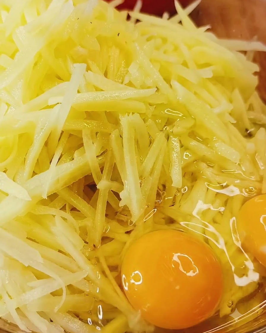 生のエディブルな千切りじゃがいもと調味料が入ったボウルに、卵を直接割り入れているところ