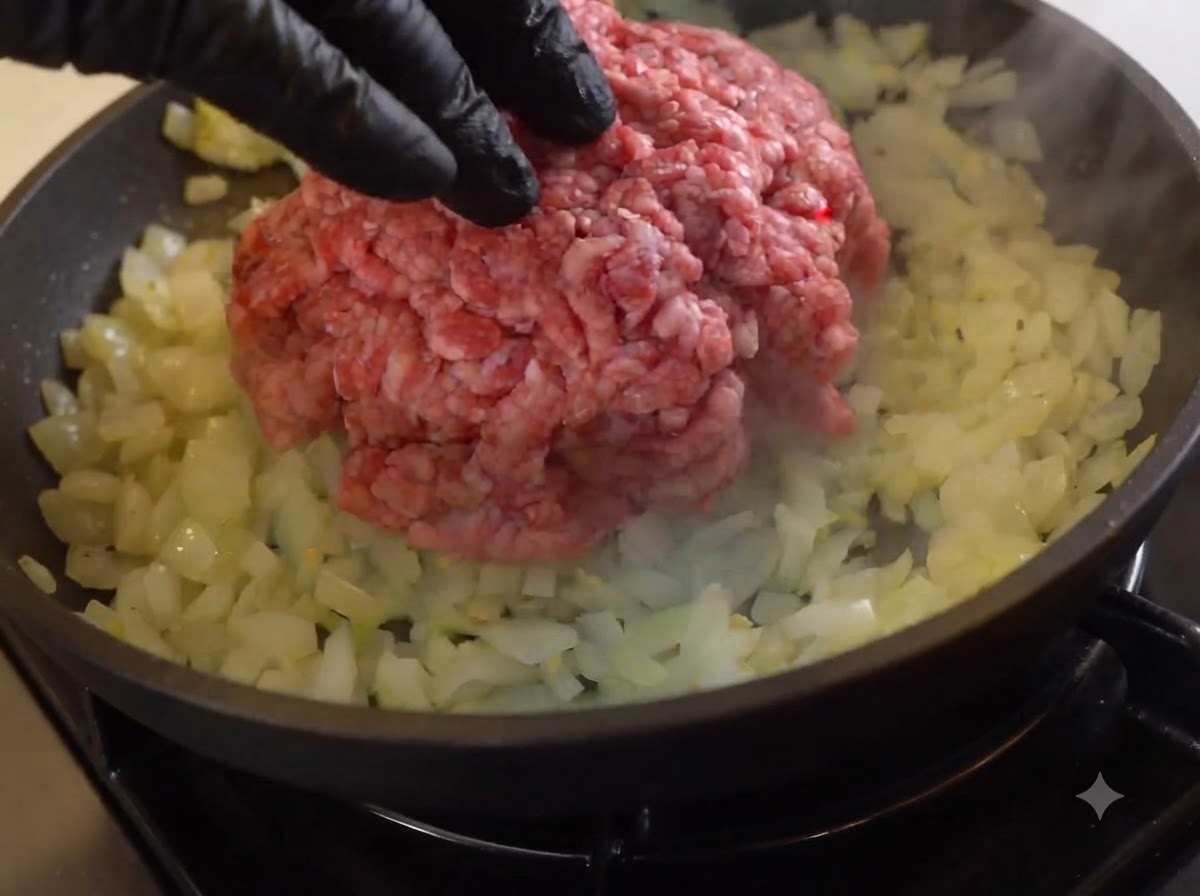 スキレットの中で、炒めたタマネギの上に生の牛ひき肉の塊が乗っている様子。
