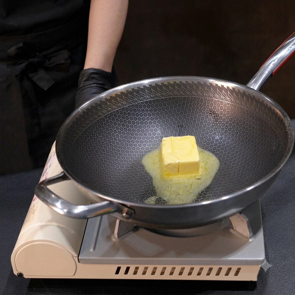 黄色いバターのブロックが金属製の中華鍋の中で溶けていき、火にかけながら小さな泡立て器で混ぜられている様子。