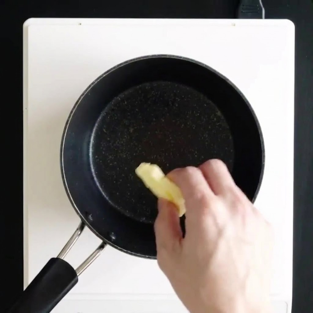 白いコンロの上の黒いソースパンに、手でバターの塊を入れて溶かしているところ。