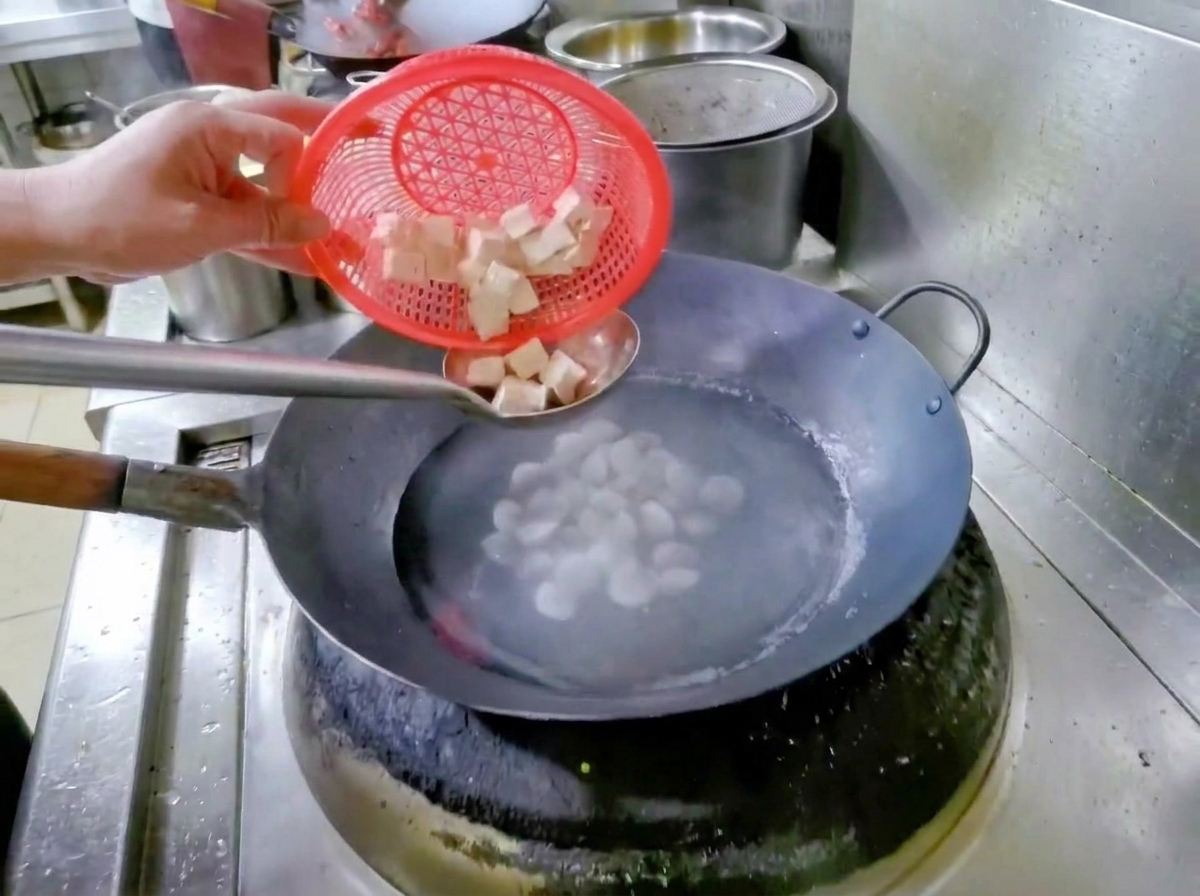 赤いザルを持った手が、水とアサリの入った鍋にさいの目切りの豆腐を入れている様子。