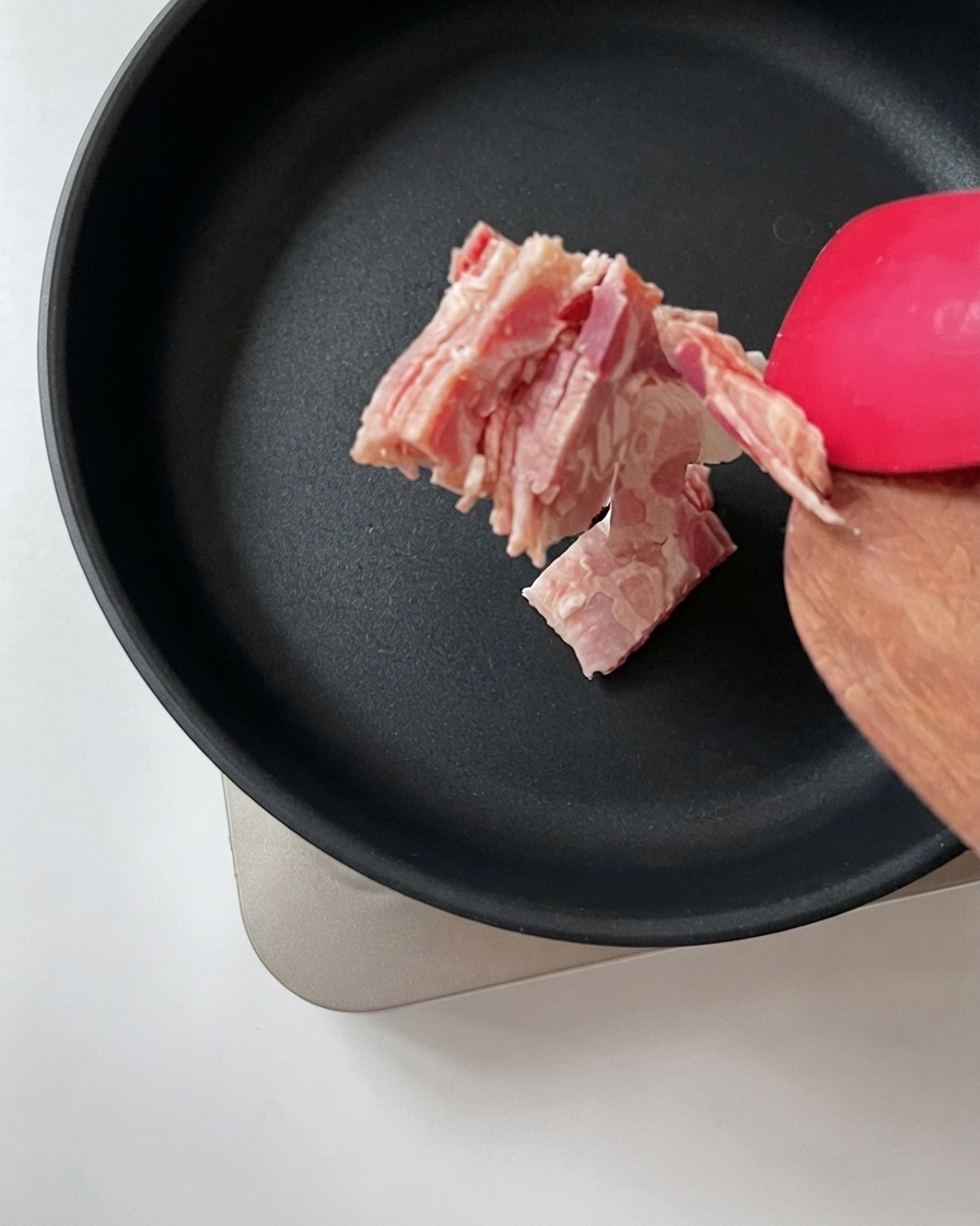 木製のスパチュラを使って、刻んだベーコンを熱した黒いフライパンに移しているところ。