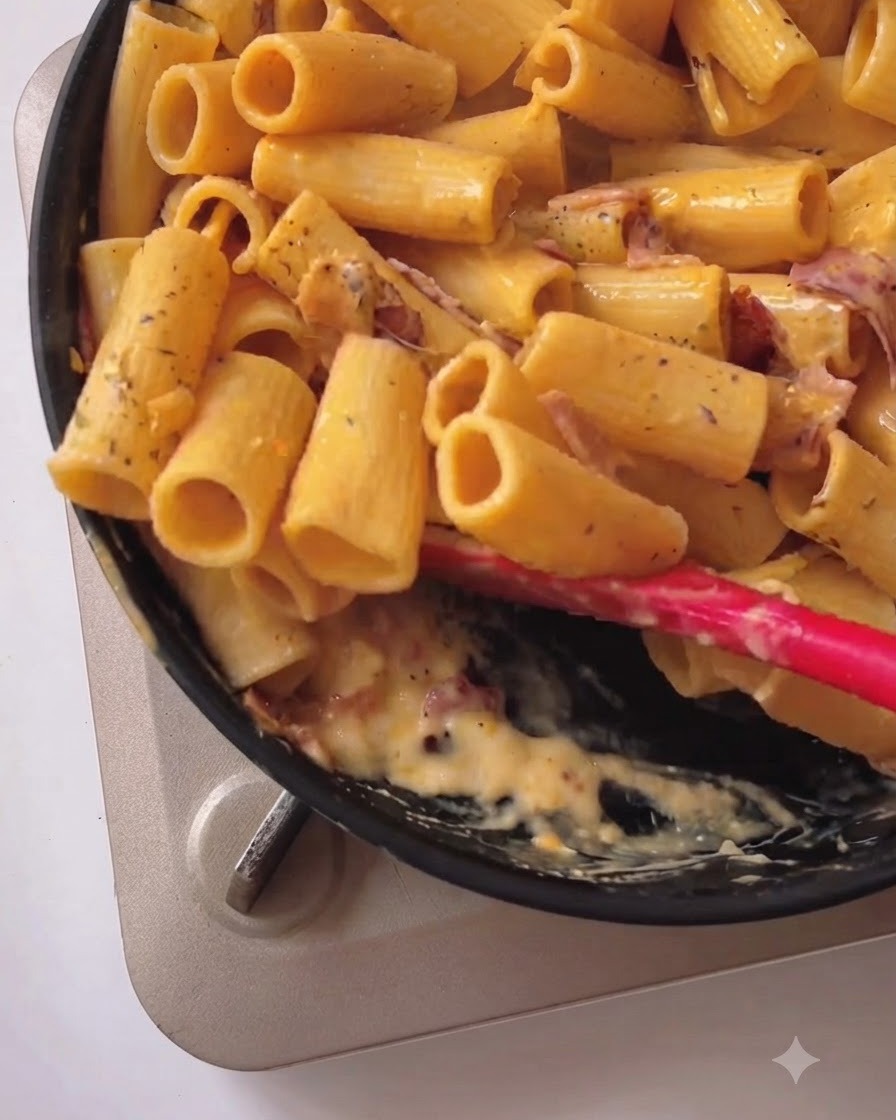 黒いフライパンの中で、赤いシリコンスパチュラを使い、リガトーニパスタとクリーミーな黄色いソース、ベーコンを和えている様子。